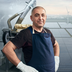 Foto von einem Mann mittleren Alters, der eine blaue Schürze trägt, vor Rohren steht und mit in die Seite gestützten Händen direkt in die Kamera schaut