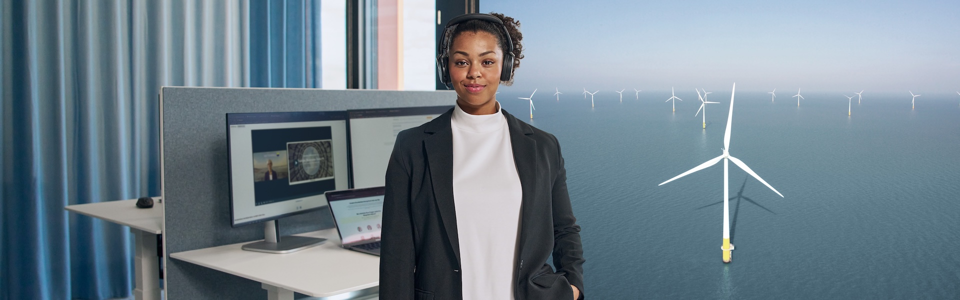 Frau schaut in die Kamera mit Kopfhörern und Bildschirm im Hintergrund.