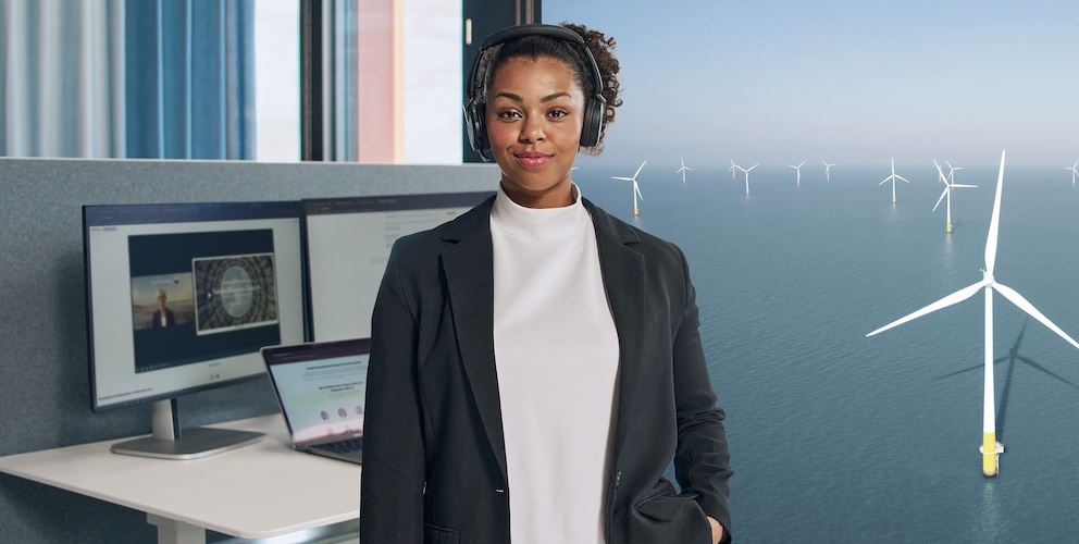 Frau schaut in die Kamera mit Kopfhörern und Bildschirm im Hintergrund.