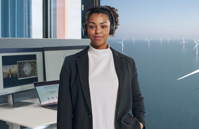 Frau schaut in die Kamera mit Kopfhörern und Bildschirm im Hintergrund.