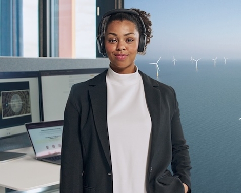 Frau schaut in die Kamera mit Kopfhörern und Bildschirm im Hintergrund.