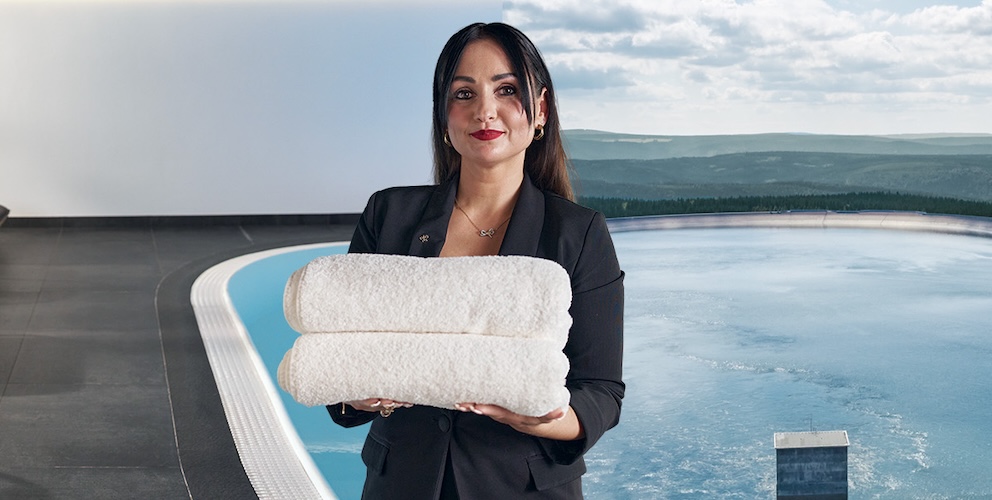 Frau hält einen Stapel mit Handtüchern. Im Hintergrund ist ein Pool mit Liegen und eine Landschaft mit Gewässer zu sehen.