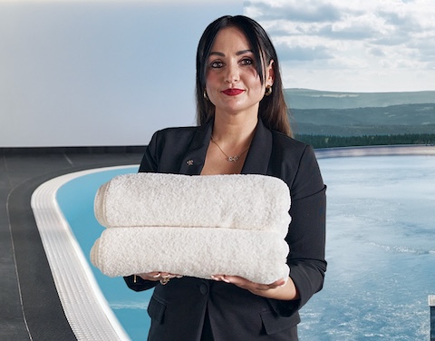 Frau hält einen Stapel mit Handtüchern. Im Hintergrund ist ein Pool mit Liegen und eine Landschaft mit Gewässer zu sehen.