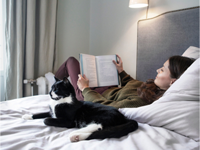 Person liegt entspannt auf einem Bett und liest ein Buch, während eine schwarz-weiße Katze am Fußende liegt.