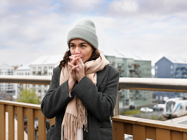 Frau steht draußen auf einer Terrasse, trägt Winterkleidung mit Mütze und Schal und wärmt sich die Hände, während im Hintergrund moderne Gebäude zu sehen sind.