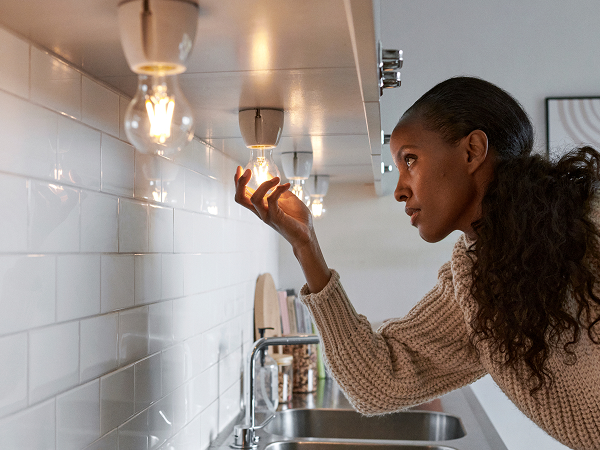 Das Bild zeigt eine Frau, die in einer modernen Küche eine Glühbirne justiert. Sie ist auf die Aufgabe konzentriert und hat einen nachdenklichen Ausdruck im Gesicht. Der Hintergrund zeigt ein sauberes, minimalistisches Design mit einer hellen, weißen Fliesenzwand und einer gut beleuchteten Umgebung. Die Kleidung der Frau wirkt lässig und bequem, was zur heimischen, entspannten Atmosphäre des Raums beiträgt.