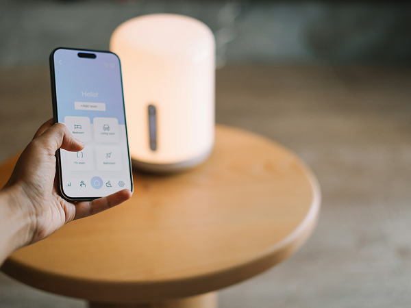 Hand hält Smartphone mit geöffneter Smart-Home-App, im Hintergrund steht eine leuchtende Lampe auf einem Holztisch – Symbol für vernetzte Haustechnik.