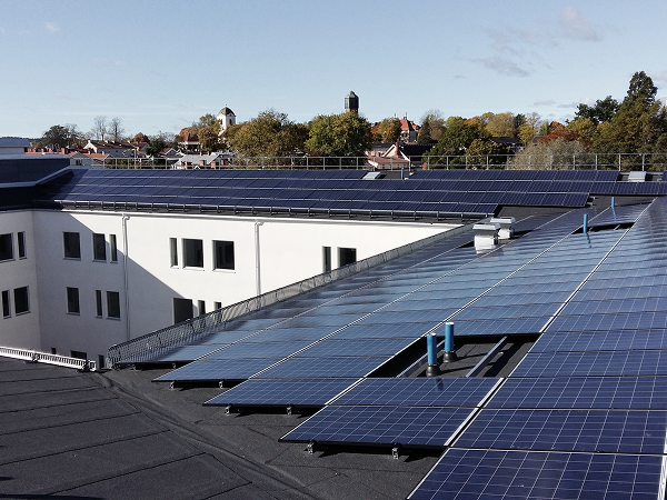 Dachfläche mit zahlreichen Solarpanels auf modernen Wohngebäuden, im Hintergrund Bäume und blauer Himmel – Beispiel für städtische Nutzung von Solarenergie.