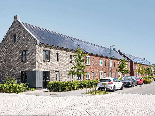 Das Bild zeigt ein modernes Wohngebäude mit Solarpanelen auf dem Dach. Die Architektur kombiniert traditionelle Stein- mit zeitgenössischen Backsteinmauern. Der Bereich vor dem Gebäude ist gepflegt, mit geparkten Autos.