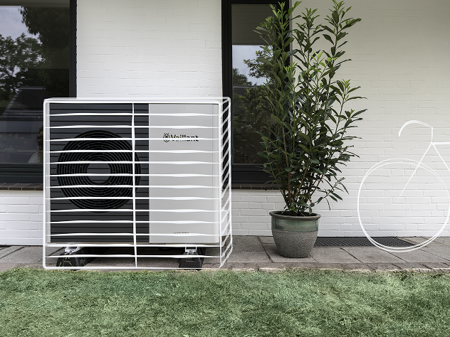 Das Bild zeigt eine Wärmepumpe, die weiß übermalt wurde, um zu demonstrieren, wie sie in die Umgebung integriert und optisch versteckt werden kann. Sie steht vor einer Wand, neben einer Pflanze und auf Rasen.