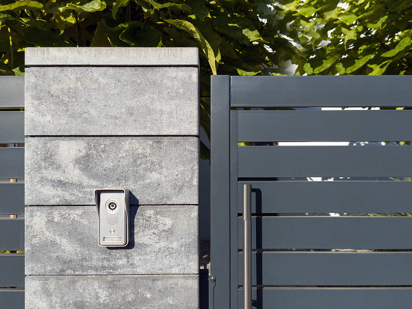 Das Bild zeigt ein modernes, graues Tor mit einem Steinsäulenpfeiler und einem einfachen Schloss. Im Hintergrund sind Pflanzen oder Bäume zu sehen.