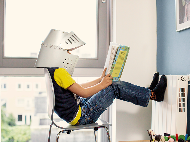 Kind sitzt entspannt auf einem Stuhl mit den Füßen auf der Heizung, liest ein Buch und trägt einen Ritterhelm.