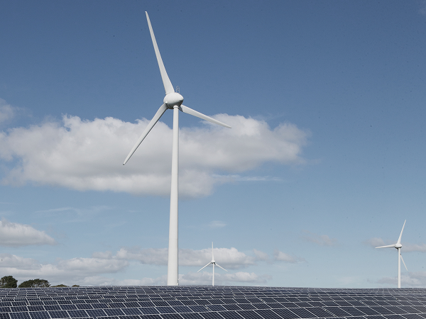 Windkraftanlage vor blauem Himmel mit weißen Wolken, im Vordergrund eine Fläche mit Solarpanels – Symbol für erneuerbare Energien.