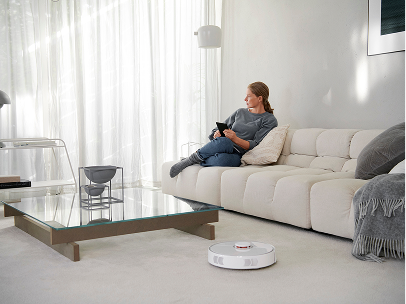 Person sitzt entspannt auf einem hellen Sofa in einem modern eingerichteten Wohnzimmer, während ein Saugroboter auf dem Boden fährt.