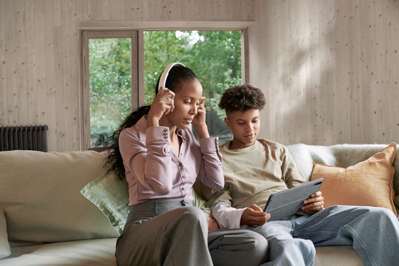 Mutter hört Musik und schaut mit Sohn auf ein Tablet, beide sitzen auf dem Sofa