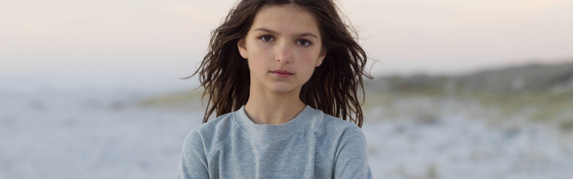 Mädchen mit braunen, langen Haaren steht an einem hellen Strand mit schemenhaften Dünen im Hintergrund und schaut direkt in die Kamera.