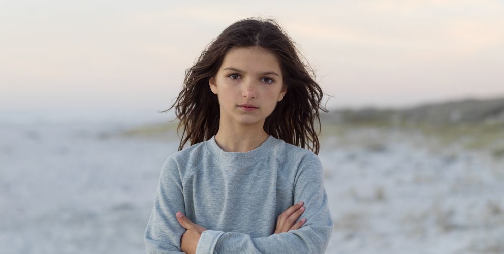 Mädchen mit braunen, langen Haaren steht an einem hellen Strand mit schemenhaften Dünen im Hintergrund und schaut direkt in die Kamera.