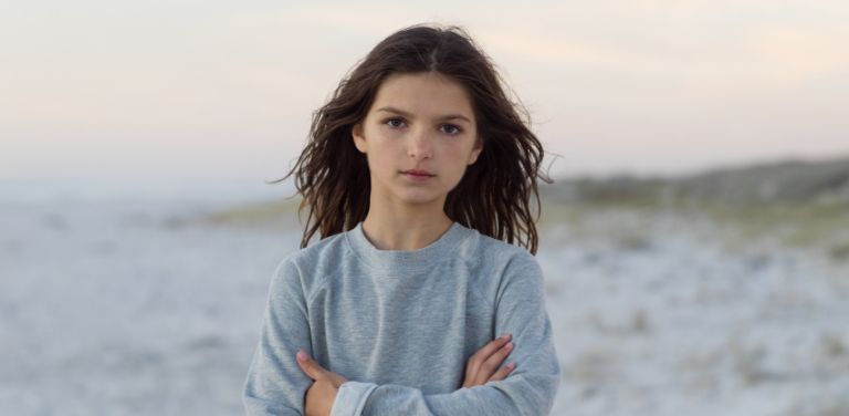 Mädchen mit braunen, langen Haaren steht an einem hellen Strand mit schemenhaften Dünen im Hintergrund und schaut direkt in die Kamera.