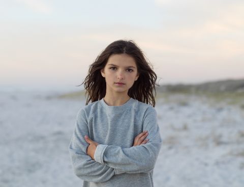 Mädchen mit braunen, langen Haaren steht an einem hellen Strand mit schemenhaften Dünen im Hintergrund und schaut direkt in die Kamera.
