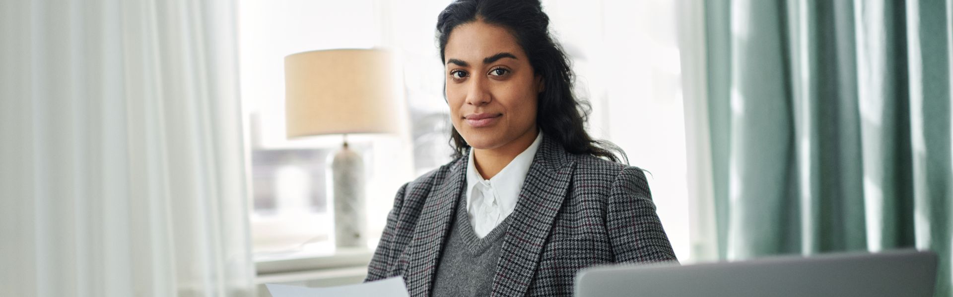 Eine Frau sitzt mit einem Papier in der Hand in einem hellen und modernen Raum und schaut lächelnd in die Kamera. Vor ihr erkennt man die Kante eines Laptops.