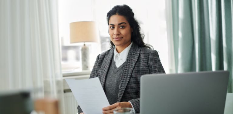 Eine Frau sitzt mit einem Papier in der Hand in einem hellen und modernen Raum und schaut lächelnd in die Kamera. Vor ihr erkennt man die Kante eines Laptops.