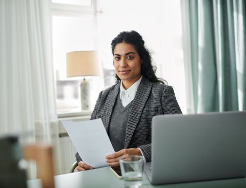 Eine Frau sitzt mit einem Papier in der Hand in einem hellen und modernen Raum und schaut lächelnd in die Kamera. Vor ihr erkennt man die Kante eines Laptops.