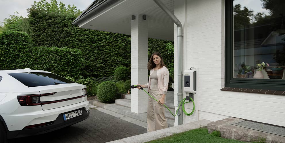 Eine Frau steht mit einem Ladekabel einer Wallbox in der Hand direkt vor ihrem Hauseingang. Im Anschitt kann man ihr weißes Elektroauto erkennen.