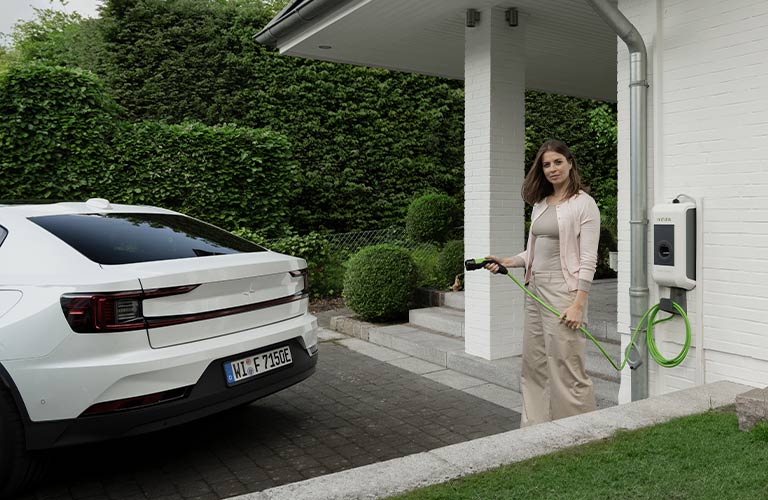 Eine Frau steht mit einem Ladekabel einer Wallbox in der Hand direkt vor ihrem Hauseingang. Im Anschitt kann man ihr weißes Elektroauto erkennen.
