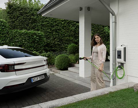 Eine Frau steht mit einem Ladekabel einer Wallbox in der Hand direkt vor ihrem Hauseingang. Im Anschitt kann man ihr weißes Elektroauto erkennen.