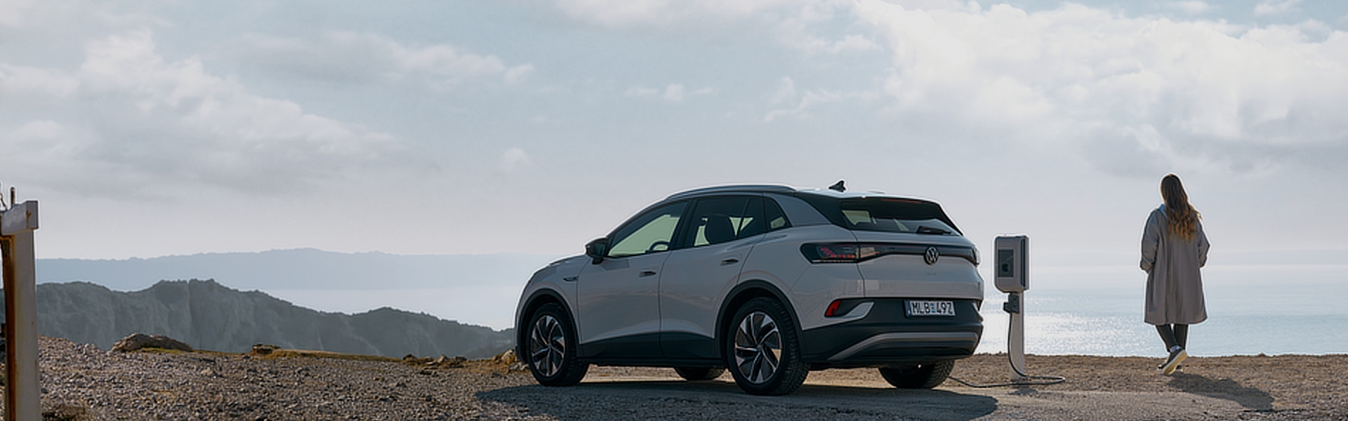 Ein weißer Elektrovolkswagen steht an einer einzelnen Ladesäule am Meer. Im Hintergrund sind Berge und das Wasser zu erkennen. Eine Person steht neben dem Auto und blickt auf das Meer.