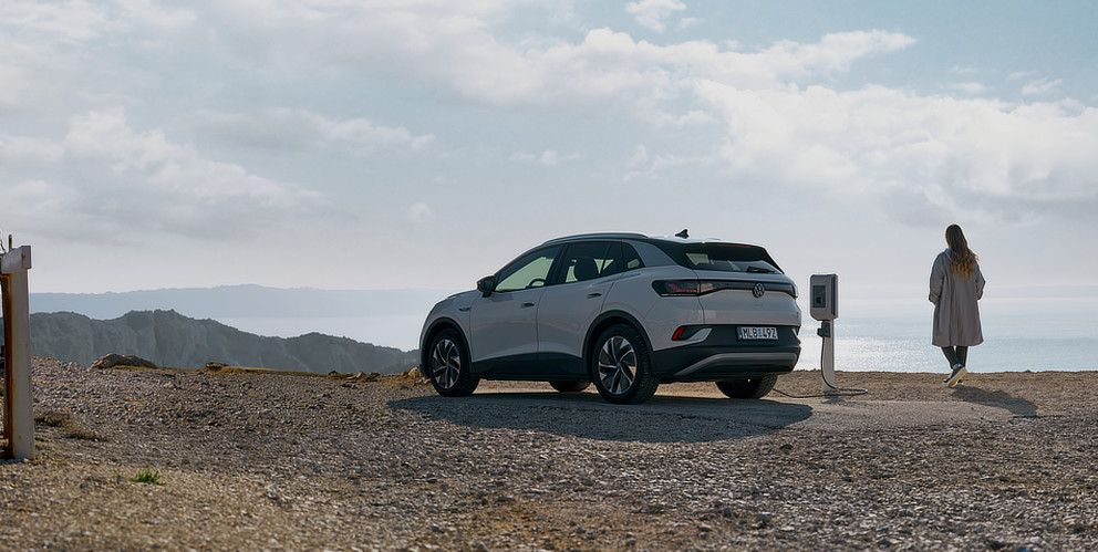 Ein weißer Elektrovolkswagen steht an einer einzelnen Ladesäule am Meer. Im Hintergrund sind Berge und das Wasser zu erkennen. Eine Person steht neben dem Auto und blickt auf das Meer.