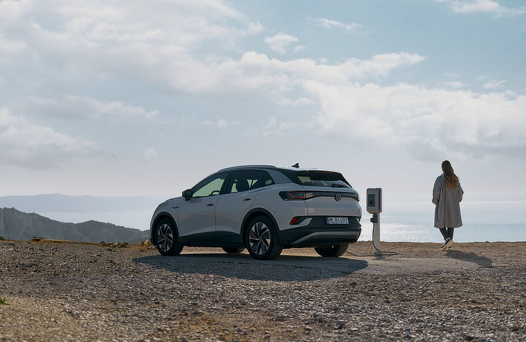 Ein weißer Elektrovolkswagen steht an einer einzelnen Ladesäule am Meer. Im Hintergrund sind Berge und das Wasser zu erkennen. Eine Person steht neben dem Auto und blickt auf das Meer.