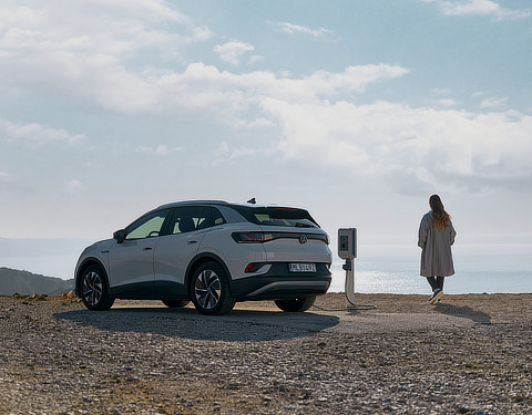 Ein weißer Elektrovolkswagen steht an einer einzelnen Ladesäule am Meer. Im Hintergrund sind Berge und das Wasser zu erkennen. Eine Person steht neben dem Auto und blickt auf das Meer.