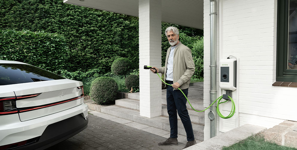 Ein Mann steht mit einem Ladekabel einer Wallbox in der Hand direkt vor seinem Hauseingang. Im bewegt sich mit dem Kabel in Richtung seines Elektroautos an der linken Bildseite.