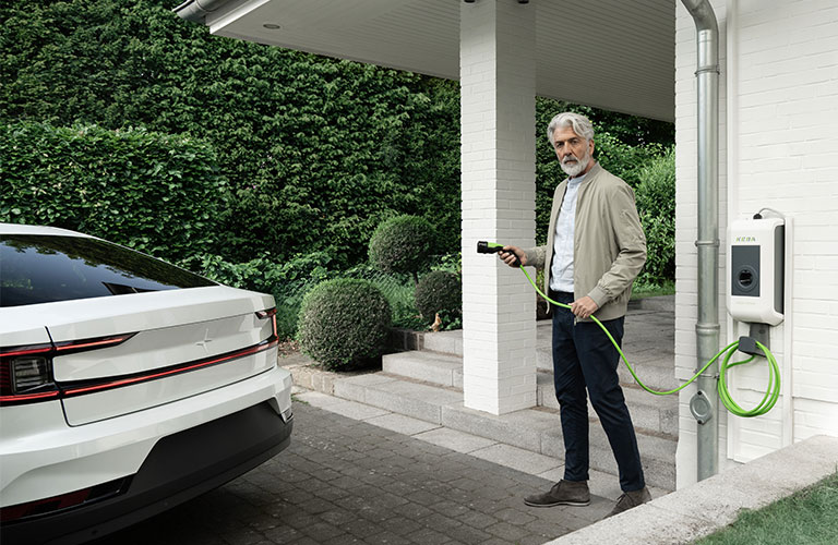 Ein Mann steht mit einem Ladekabel einer Wallbox in der Hand direkt vor seinem Hauseingang. Im bewegt sich mit dem Kabel in Richtung seines Elektroautos an der linken Bildseite.