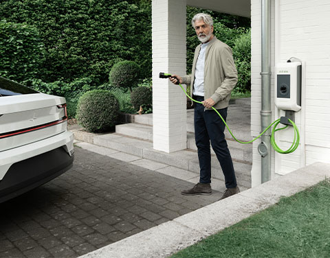 Ein Mann steht mit einem Ladekabel einer Wallbox in der Hand direkt vor seinem Hauseingang. Im bewegt sich mit dem Kabel in Richtung seines Elektroautos an der linken Bildseite.