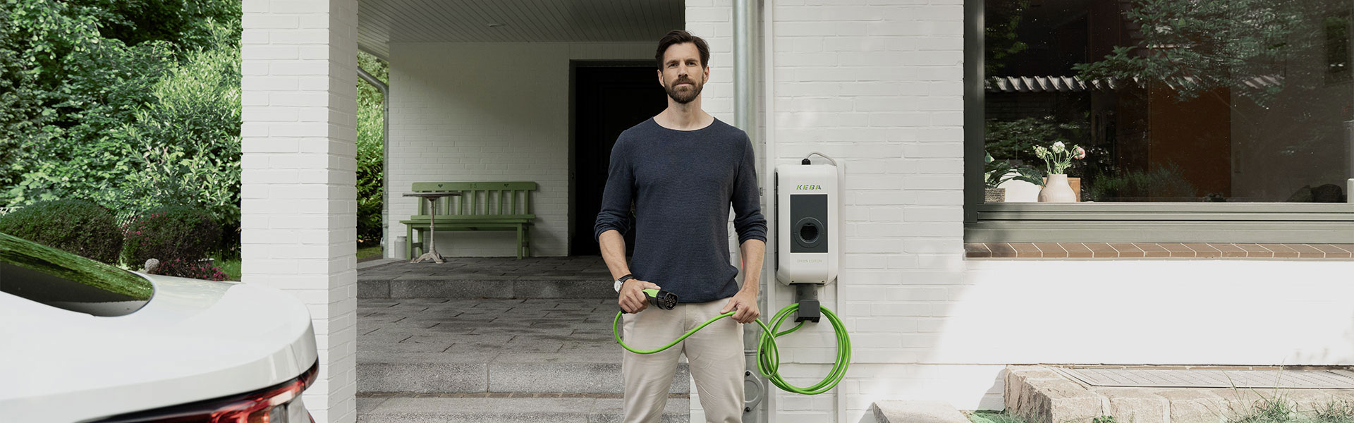 Ein Mann steht mit einem Ladekabel einer Wallbox in der Hand direkt vor seinem Hauseingang. Im Anschitt kann man ein Auto erkennen.