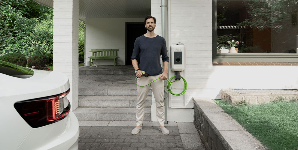 Ein Mann steht mit einem Ladekabel einer Wallbox in der Hand direkt vor seinem Hauseingang. Im Anschitt kann man ein Auto erkennen.