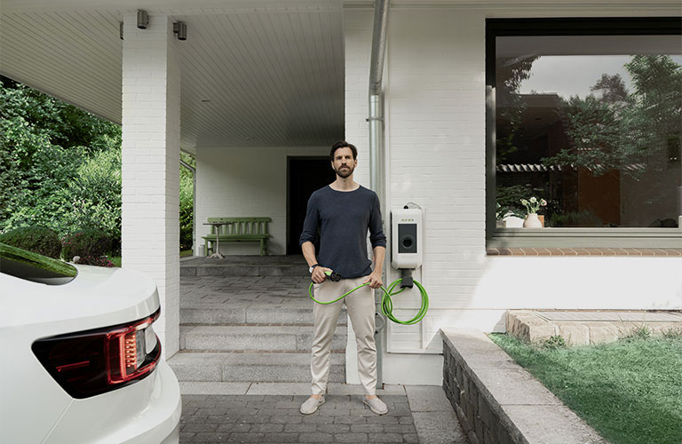 Ein Mann steht mit einem Ladekabel einer Wallbox in der Hand direkt vor seinem Hauseingang. Im Anschitt kann man ein Auto erkennen.