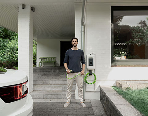 Ein Mann steht mit einem Ladekabel einer Wallbox in der Hand direkt vor seinem Hauseingang. Im Anschitt kann man ein Auto erkennen.
