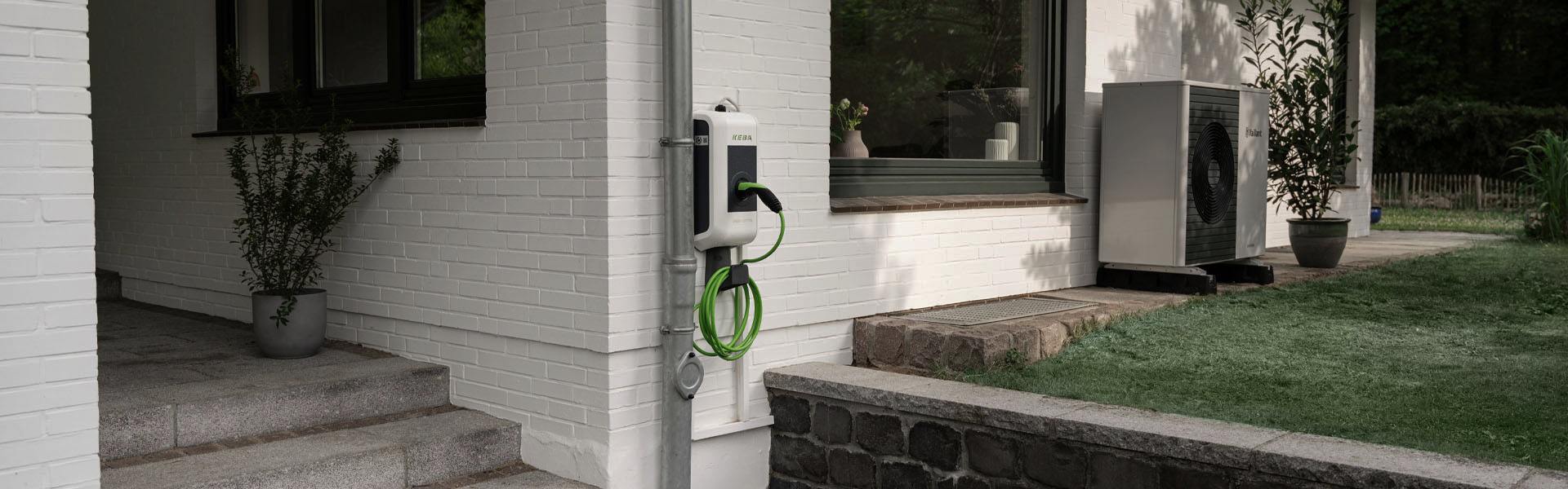 KEBA-Wallbox an einer Hauswand eines Einfamilienhauses. Im Hintergrund steht eine Wärmepumpe.