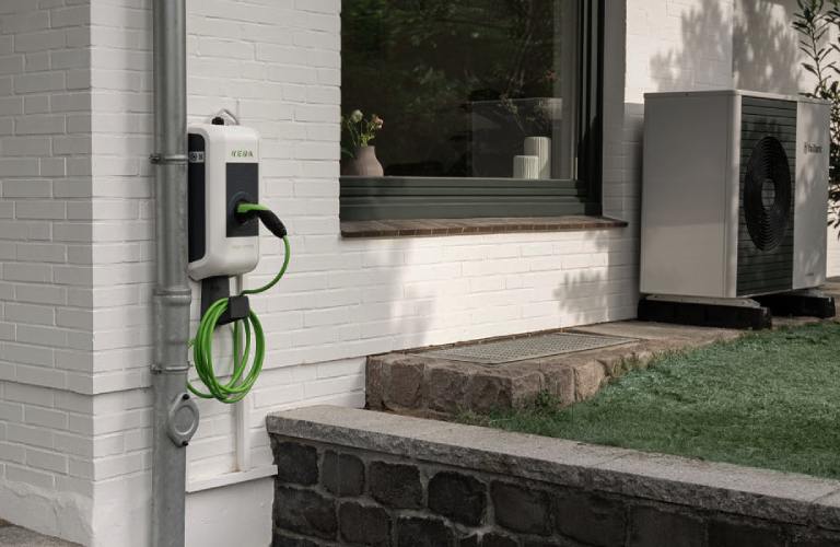 KEBA-Wallbox an einer Hauswand eines Einfamilienhauses. Im Hintergrund steht eine Wärmepumpe.