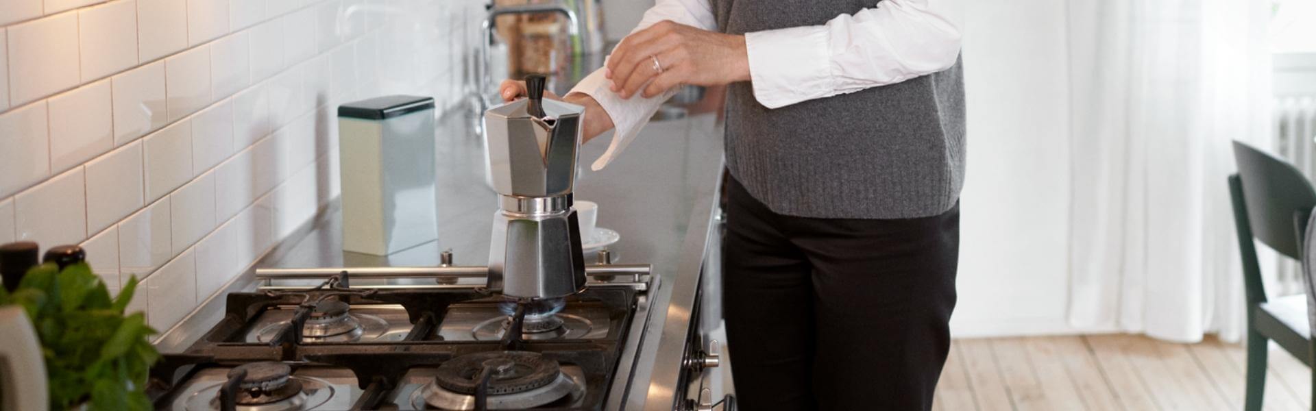 Person steht in einer modernen, hellen Küche an einem Gasherd. Sie kocht sich einen Kaffee in einem Espressokocher aus Edelstahl.