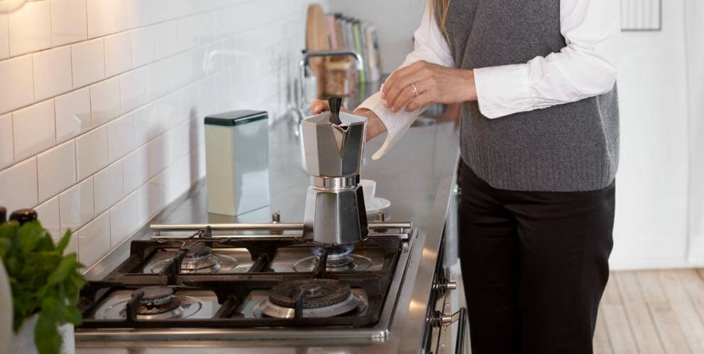 Person steht in einer modernen, hellen Küche an einem Gasherd. Sie kocht sich einen Kaffee in einem Espressokocher aus Edelstahl.