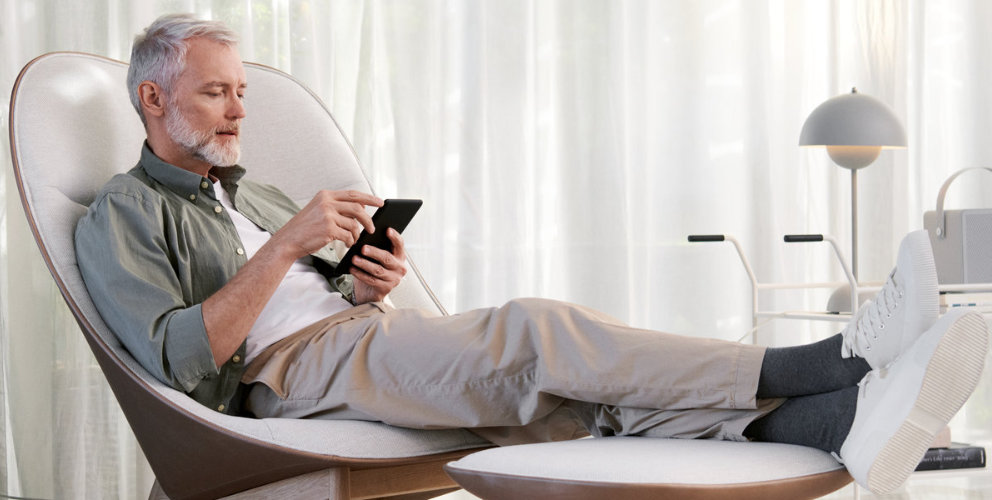 Mann sitzt im Wohnzimmer auf einem Sessel und schaut auf sein Smartphone.