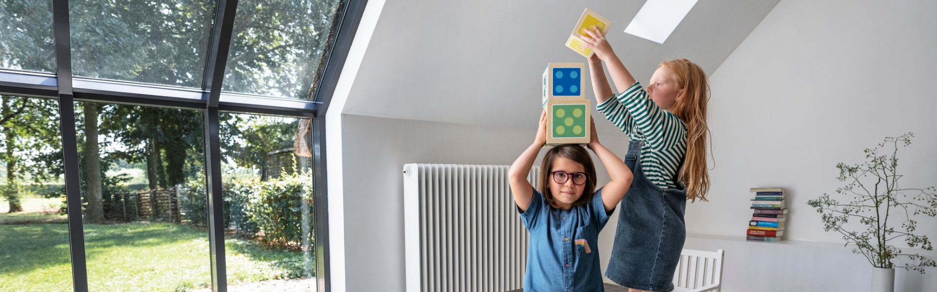 Zwei Kinder spielen mit bunten Blöcken in einem hellen Raum mit einem großen Fenster. An der Wand ist ein großer Heizkörper befestigt.