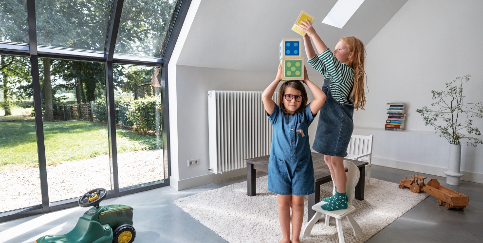 Zwei Kinder spielen mit bunten Blöcken in einem hellen Raum mit einem großen Fenster. An der Wand ist ein großer Heizkörper befestigt.