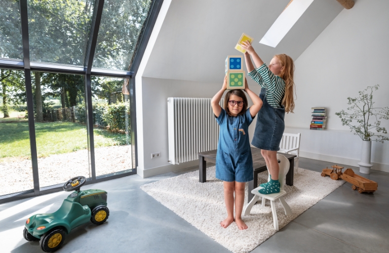 Zwei Kinder spielen mit bunten Blöcken in einem hellen Raum mit einem großen Fenster. An der Wand ist ein großer Heizkörper befestigt.