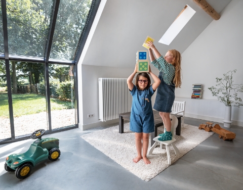 Zwei Kinder spielen mit bunten Blöcken in einem hellen Raum mit einem großen Fenster. An der Wand ist ein großer Heizkörper befestigt.