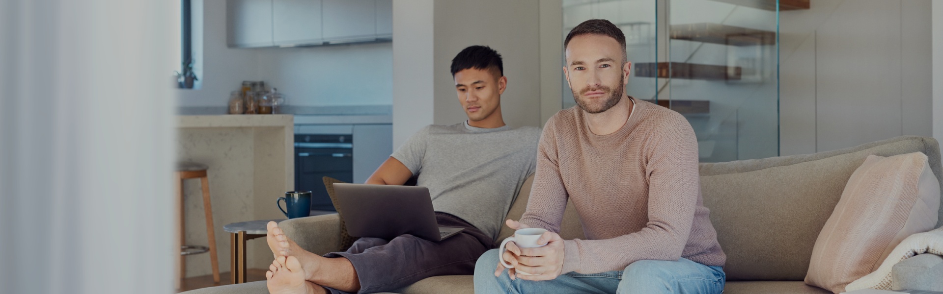 Zwei Männer sitzen zu Hause auf einem Sofa. Einer hat einen Laptop auf dem Schoß, der andere schaut freundlich in die Kamera.